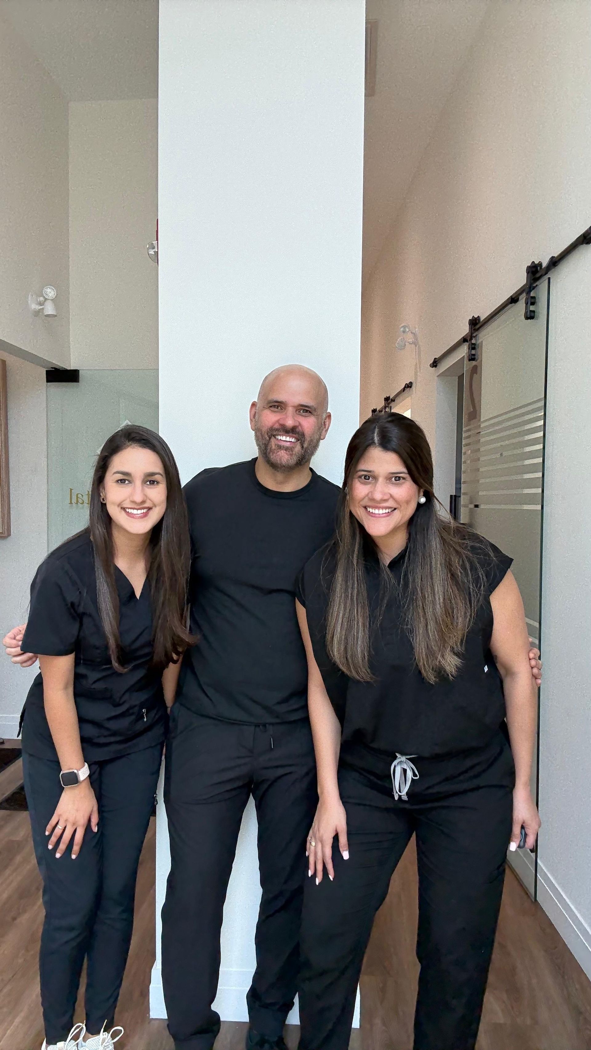 Three people smiling for a photo in an office setting. Two women in black scrubs flank a man in a black shirt and pants.
