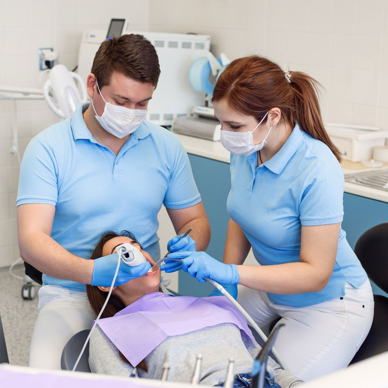 Dentist and assistant treating patient in dental office. Patient reclined, mouth open. Blue scrubs, masks, tools.