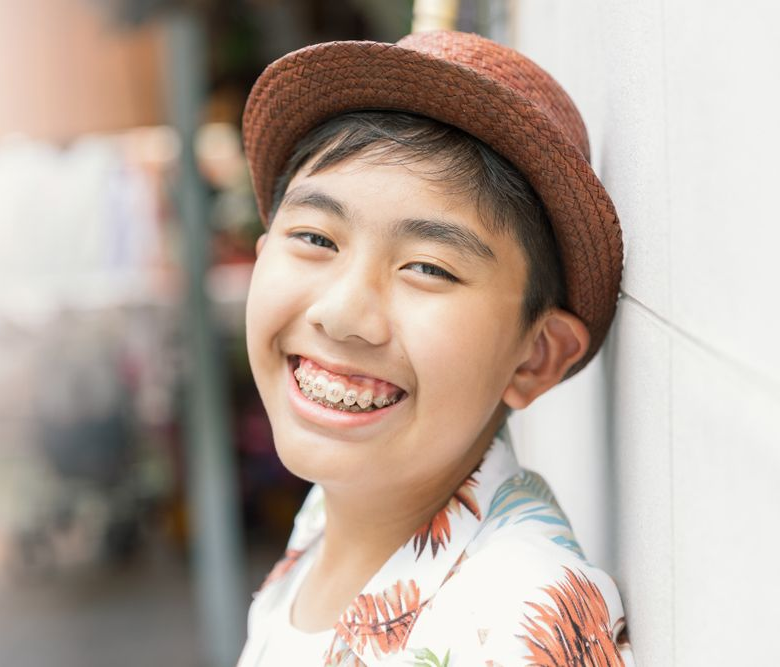 Smiling child in a brown hat leaning against a wall, wearing a floral shirt and braces
