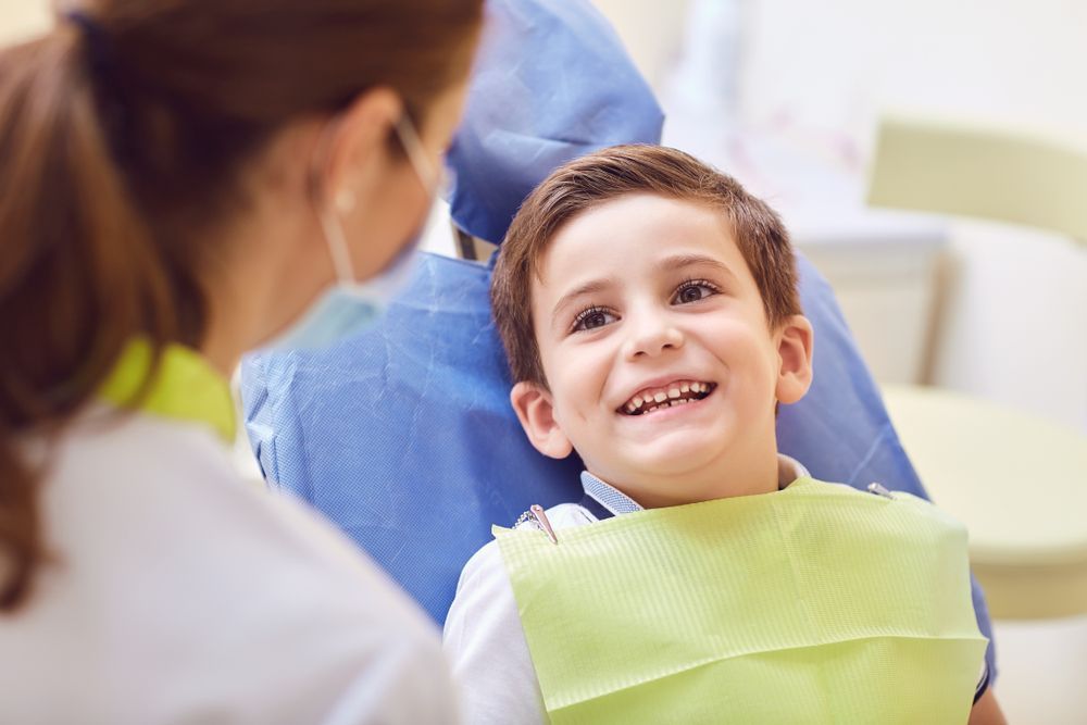 Boy smiling at a dentist, wearing a protective bib. Dentist in mask is leaning in.