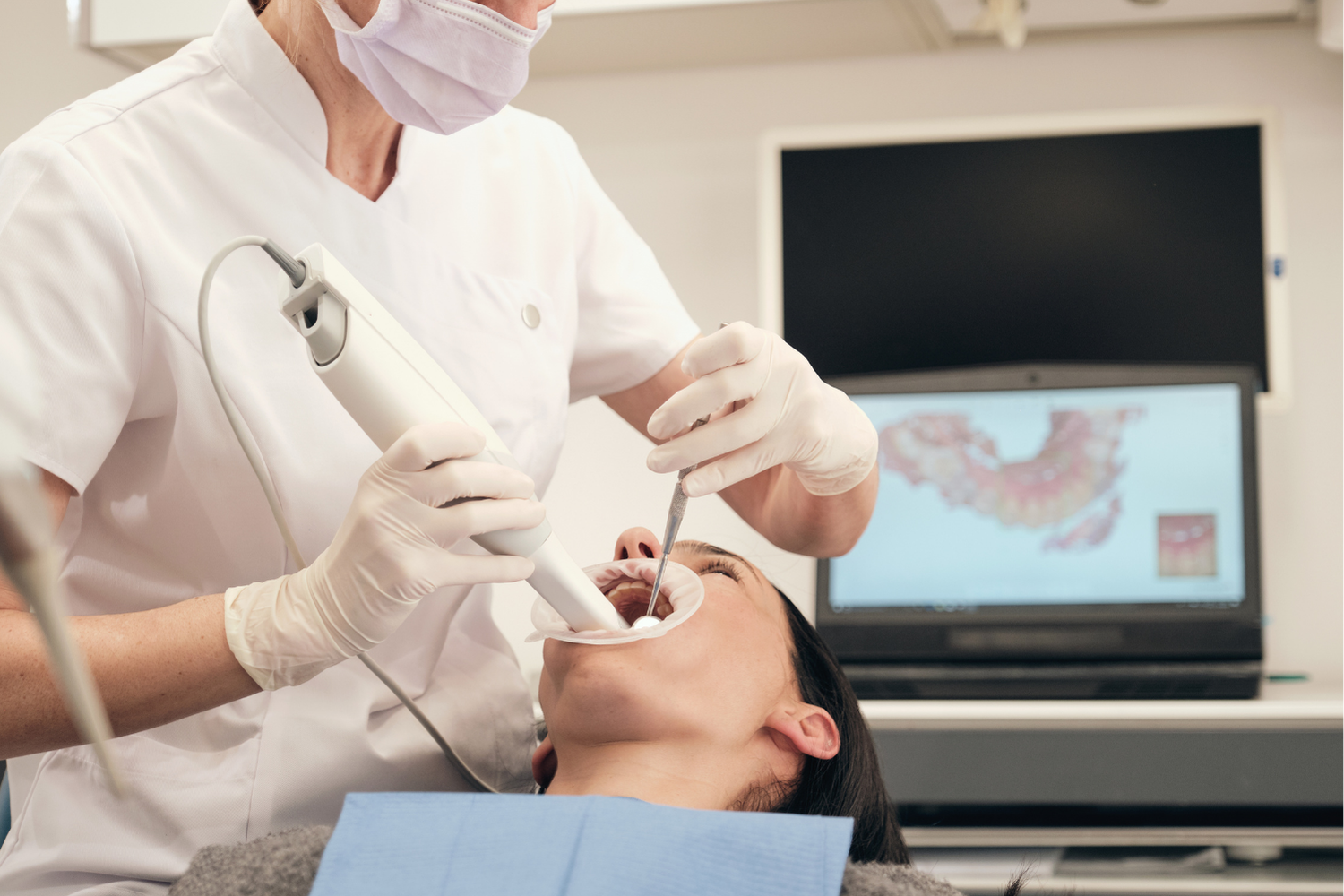 Dentist scanning patient's mouth with a digital scanner. White coat, gloves, monitor with tooth scan.