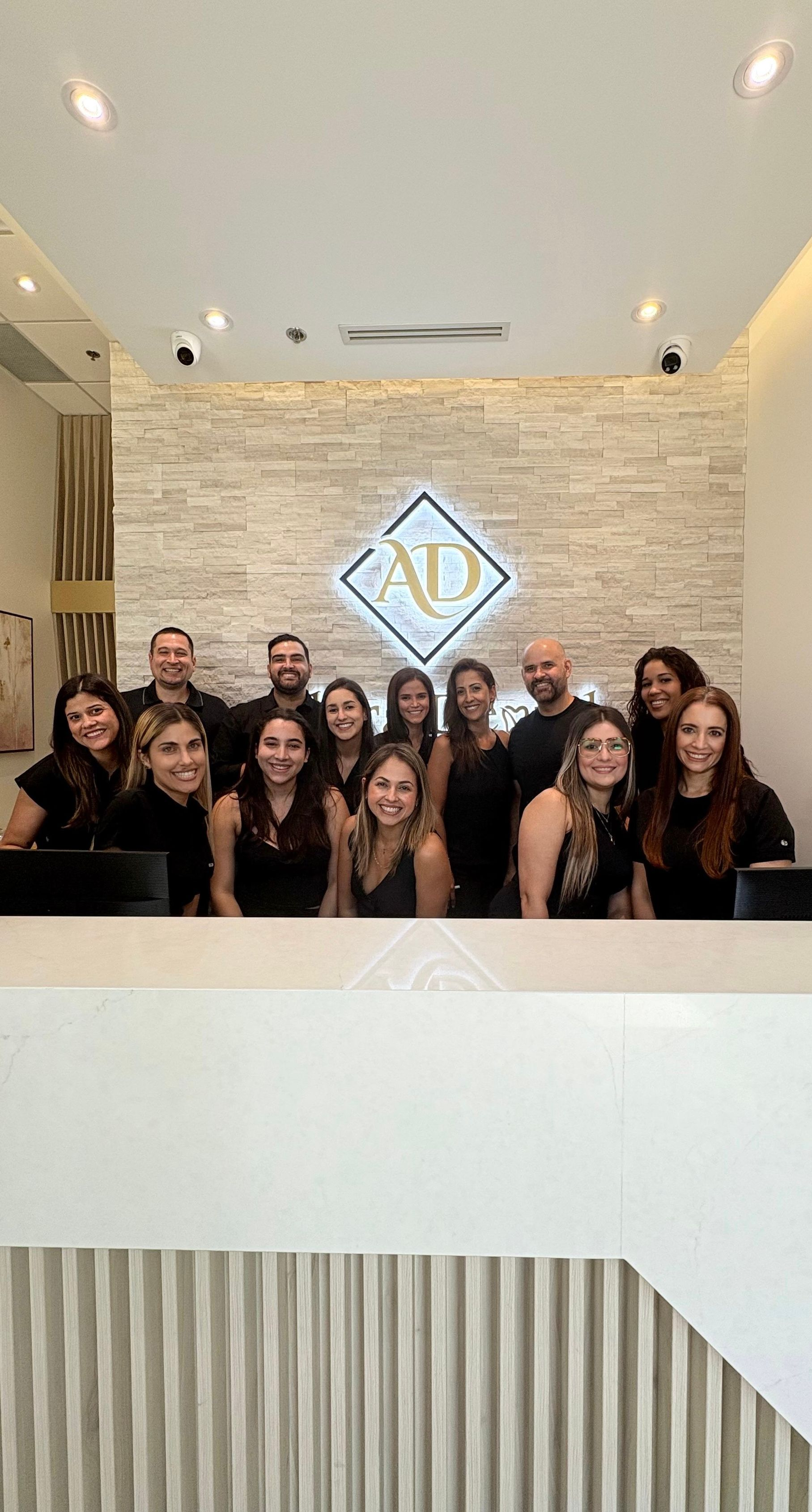 A group of people behind a white reception desk, posing in front of a dental office logo on a stone wall.