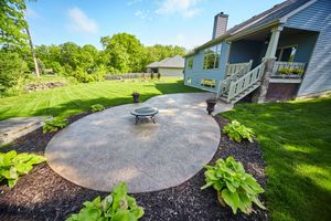 Backyard patio with concrete fire pit, green lawn, landscaped beds, and a house porch in bright sunlight