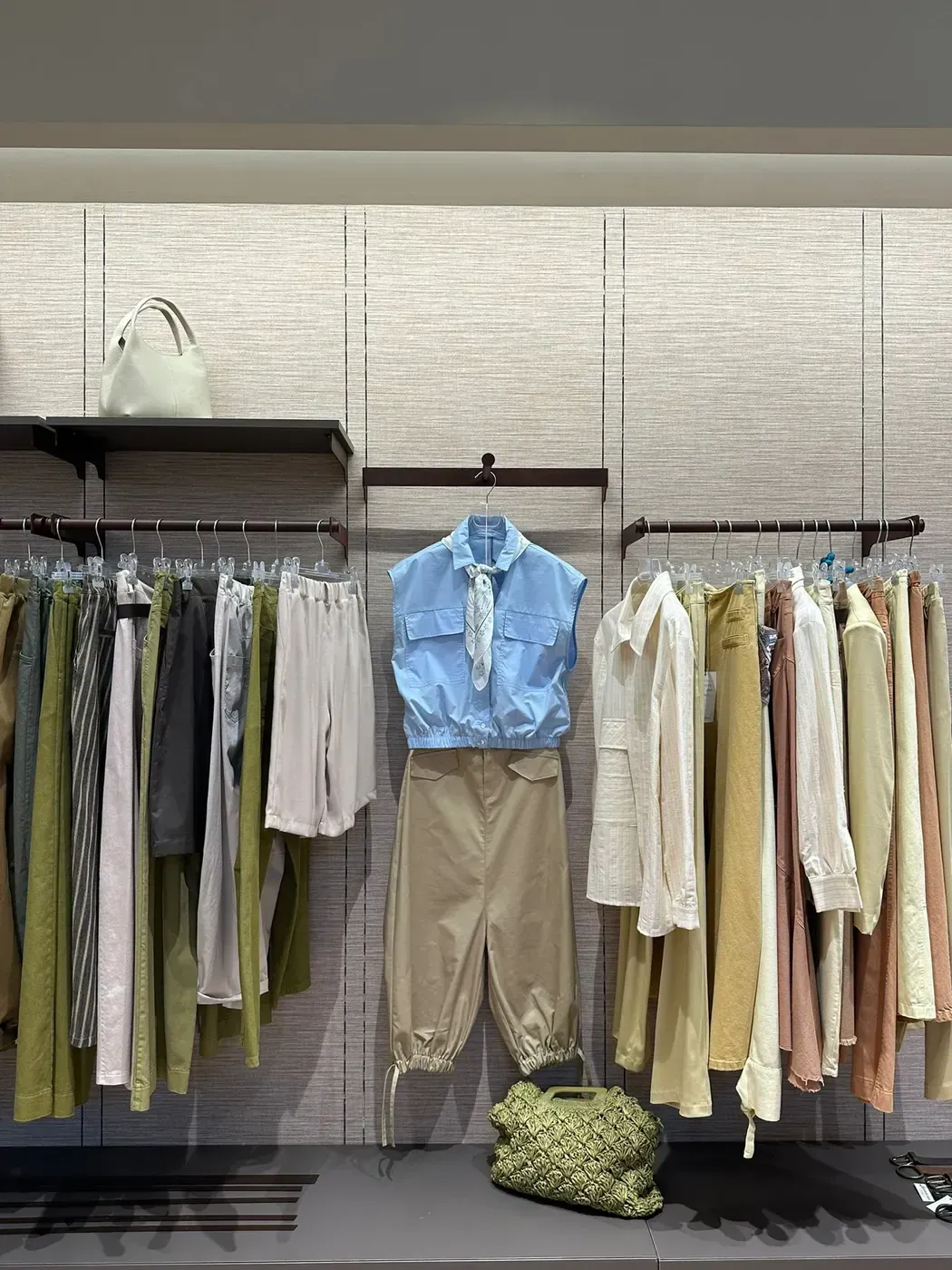 Esposizione di abiti su appendiabiti con top color pastello, pantaloni neutri e una camicia blu posizionata al centro contro una parete con texture.