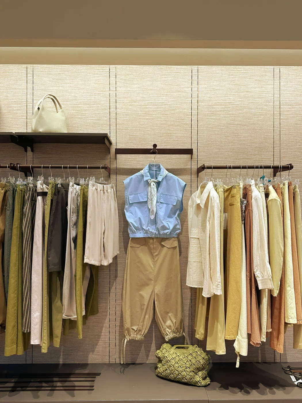 Esposizione di abiti su appendiabiti con top color pastello, pantaloni neutri e una camicia blu posizionata al centro contro una parete con texture.