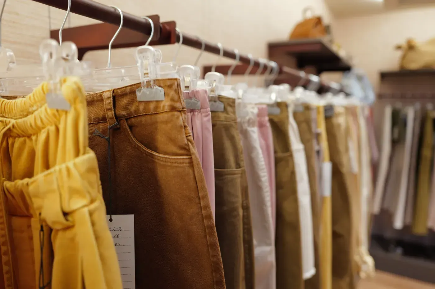 In un negozio di abbigliamento, uno scaffale con pantaloni ordinatamente appesi nei colori giallo, marrone, rosa e beige.