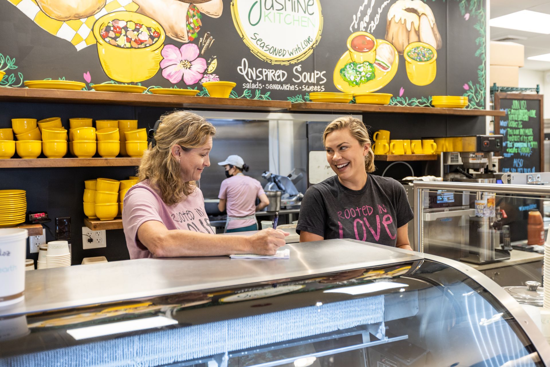 Two café workers behind a glass counter with yellow cups and a colorful menu board overhead