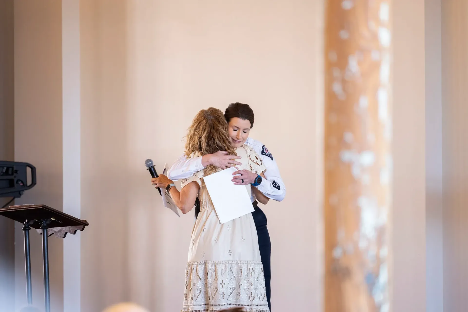 Two people embracing on a stage, one holding a microphone and papers in a bright indoor venue
