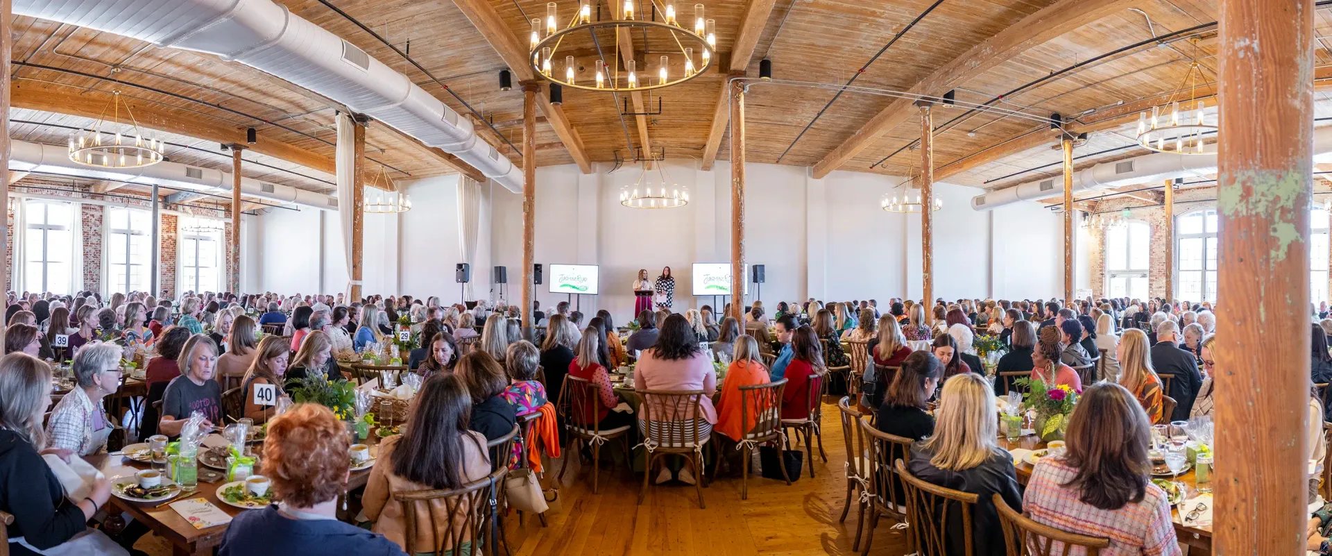Crowded wooden hall with a large seated audience facing a bright stage in front.