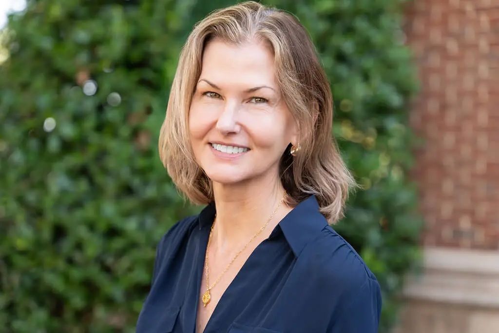 Smiling woman in a navy blouse standing outdoors with greenery and a brick wall behind her