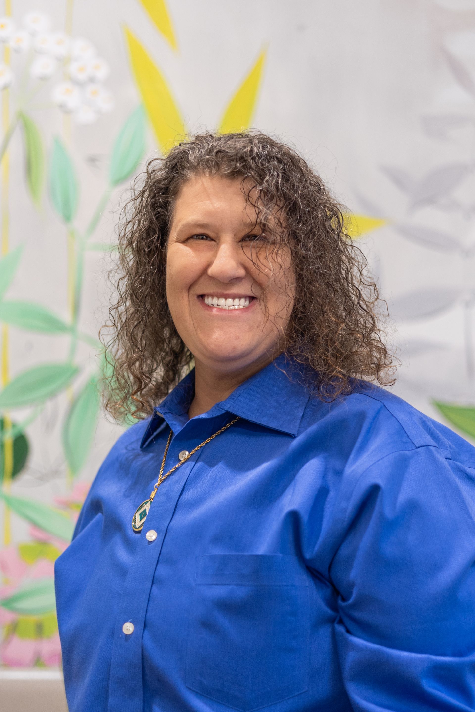 Smiling woman in a blue shirt standing before a light floral backdrop