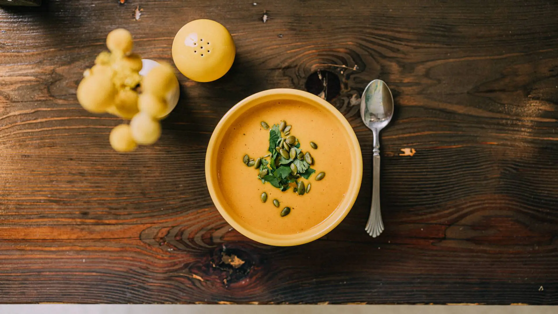 Creamy orange soup in a bowl with herbs, beside a spoon, pepper shaker, and bread on a wooden table