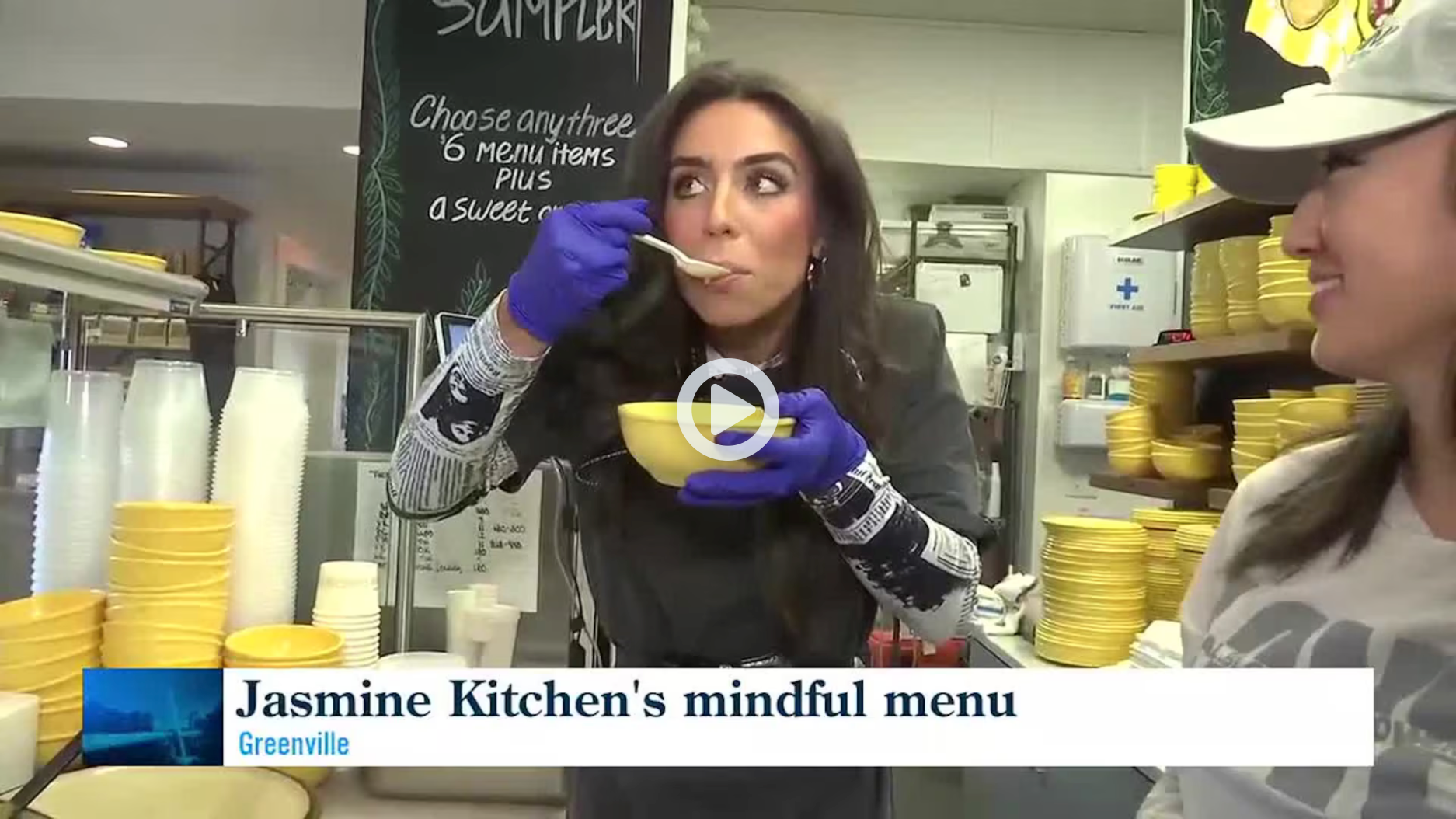 Woman sampling food at Jasmine Kitchen counter, with menu sign and jars in the background.