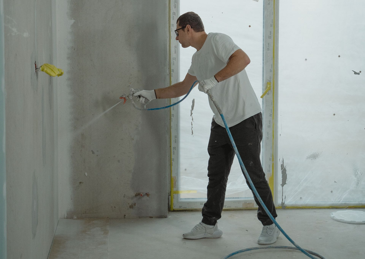 Person using a paint sprayer to coat a wall in a room under renovation