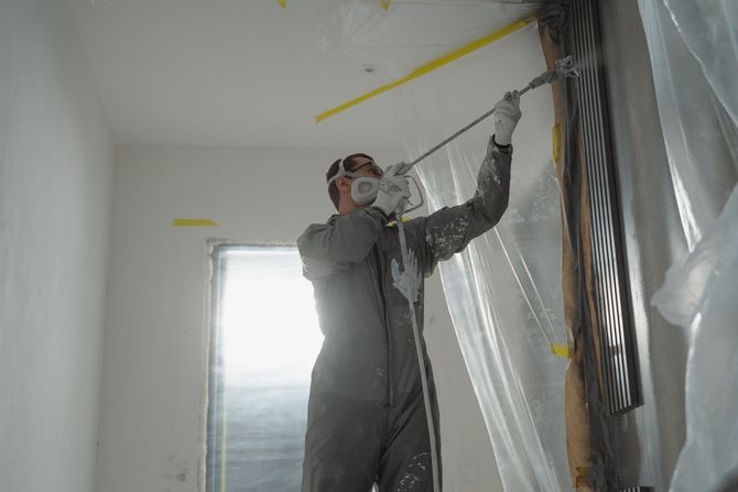 Worker in gray coveralls spray-painting a white room near a window and plastic-covered wall.