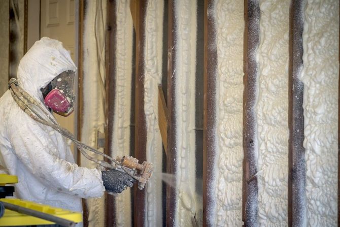 Person in protective gear spray-insulating a wooden surface.