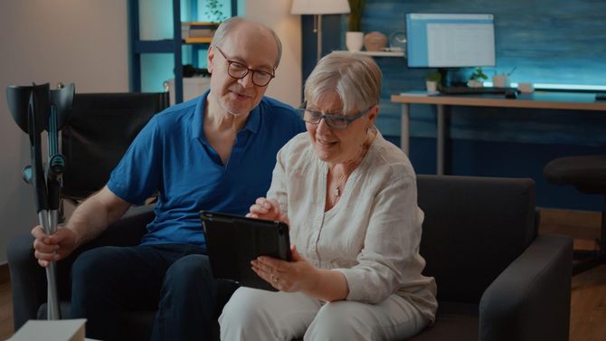 Elderly couple on a couch, looking at a tablet together. Man holds a cane. Interior setting.