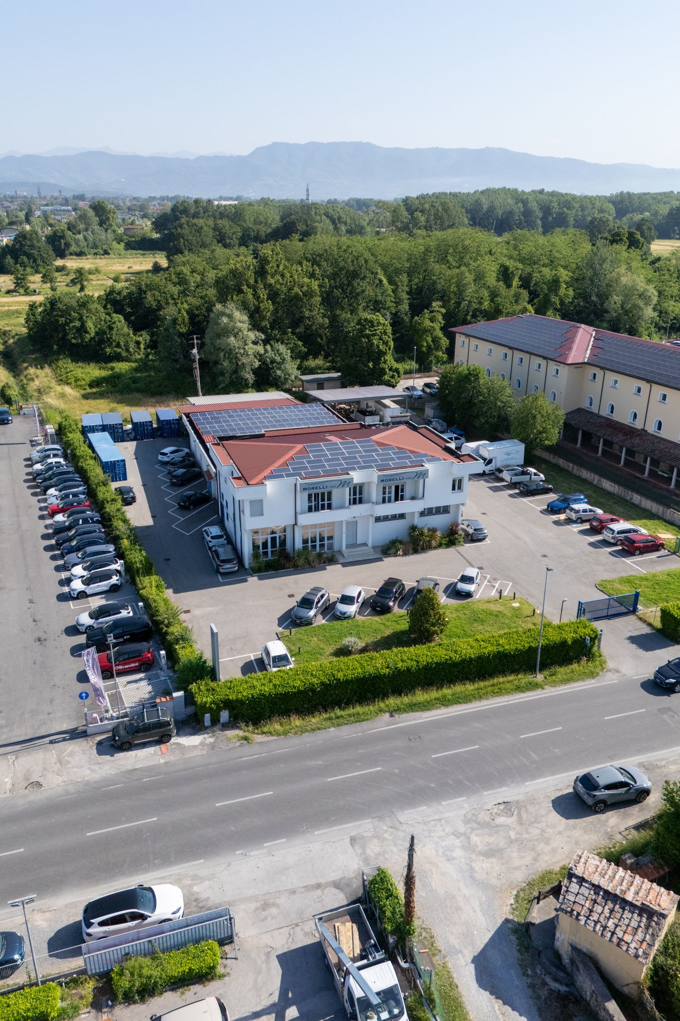 Vista aerea di un edificio bianco con il tetto rosso, accanto a un parcheggio e a una strada alberata.