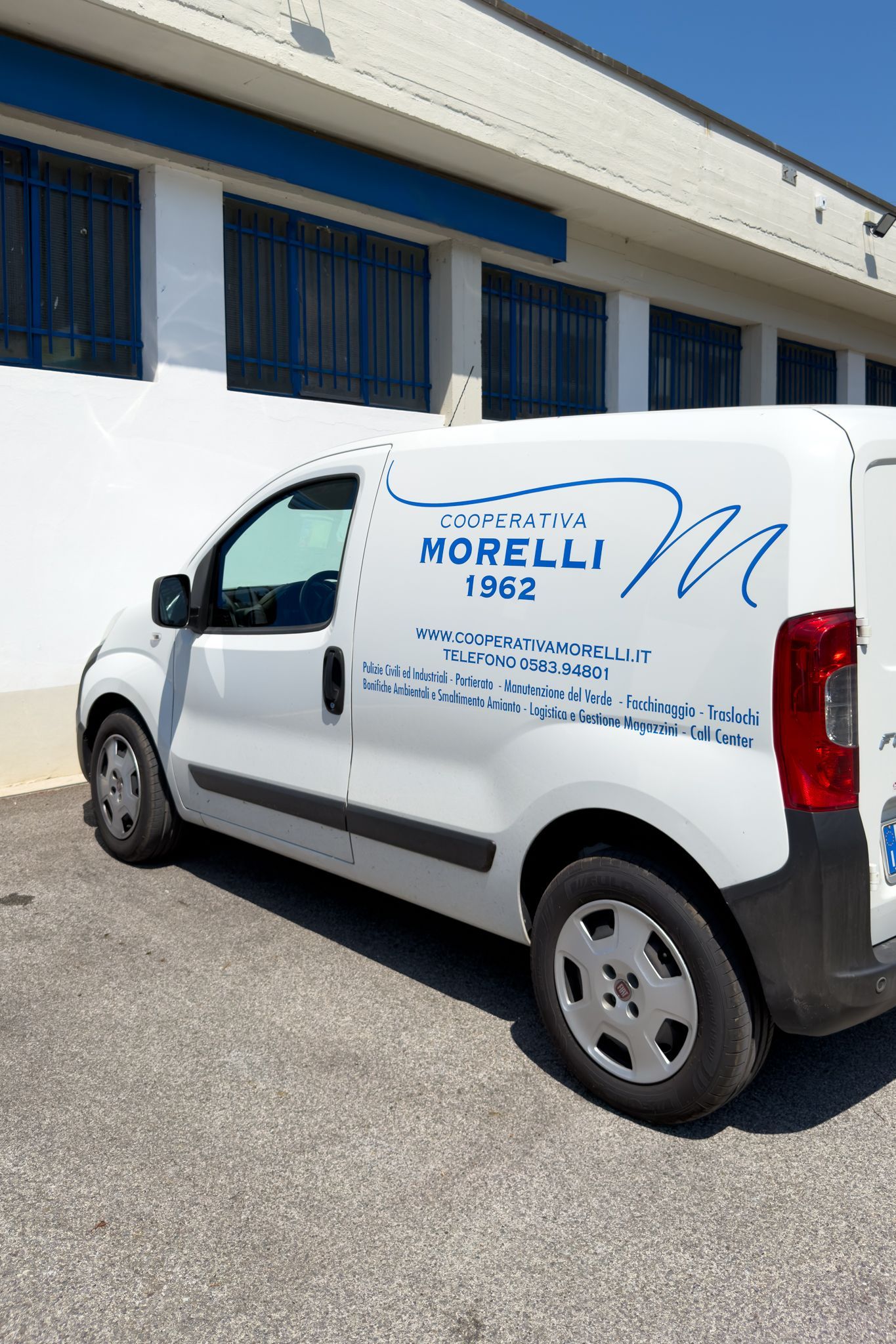 Furgone bianco per le consegne con logo aziendale blu parcheggiato accanto a un edificio con finestre blu.