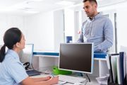 Due colleghi discutono seduti a una scrivania davanti a un monitor in un ufficio luminoso.