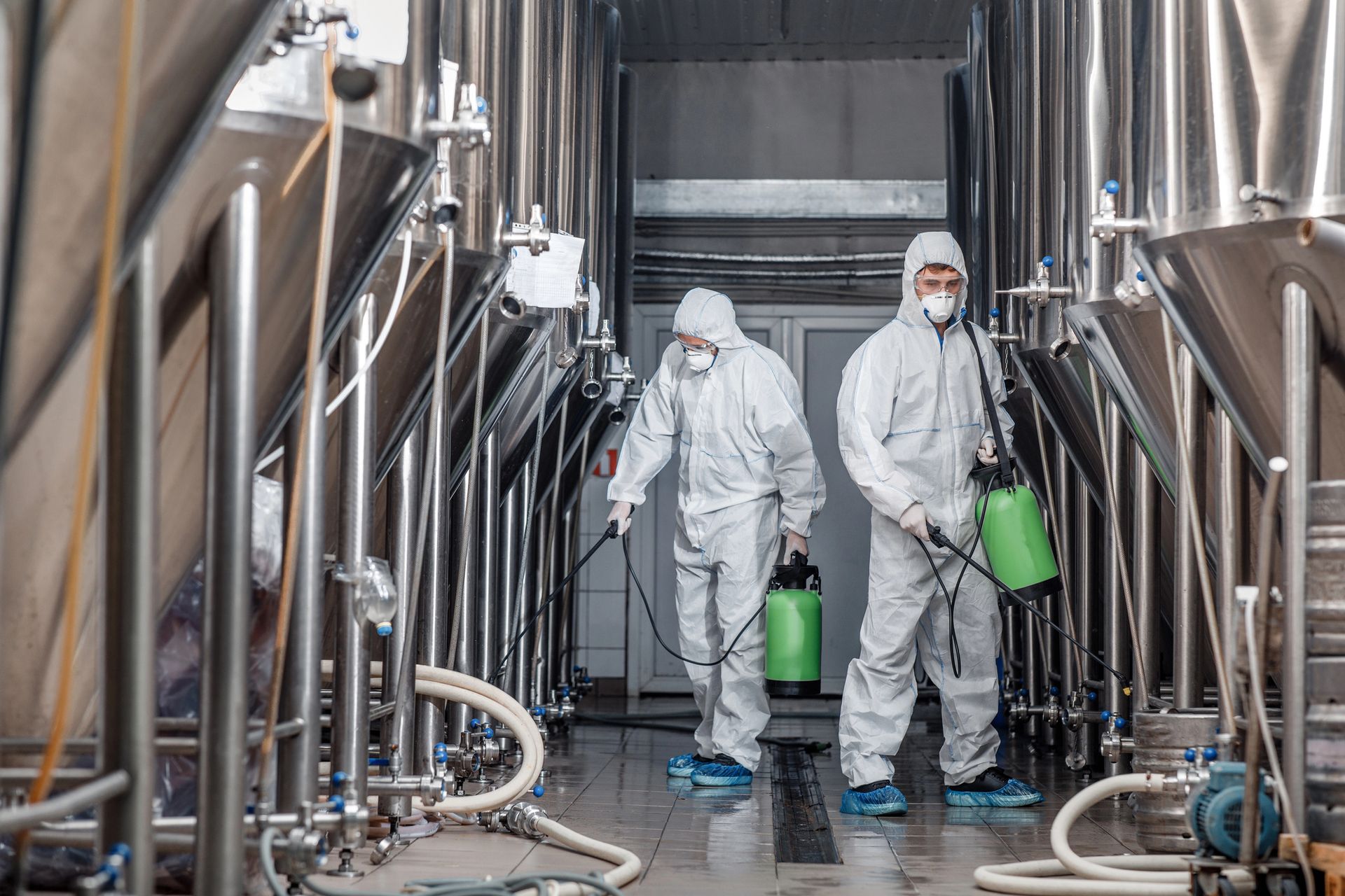 Two workers in white hazmat suits carry green containers inside an industrial processing facility.