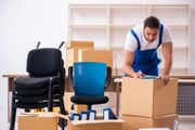 Un traslocatore in tuta blu apre una scatola di cartone accanto a una pila di sedie e a una libreria.