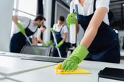 Persone con il grembiule che puliscono i tavoli in un ufficio con guanti verdi e panni gialli.