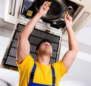 Tecnico con camicia gialla e tuta blu che ripara un'unità di ventilazione a soffitto.