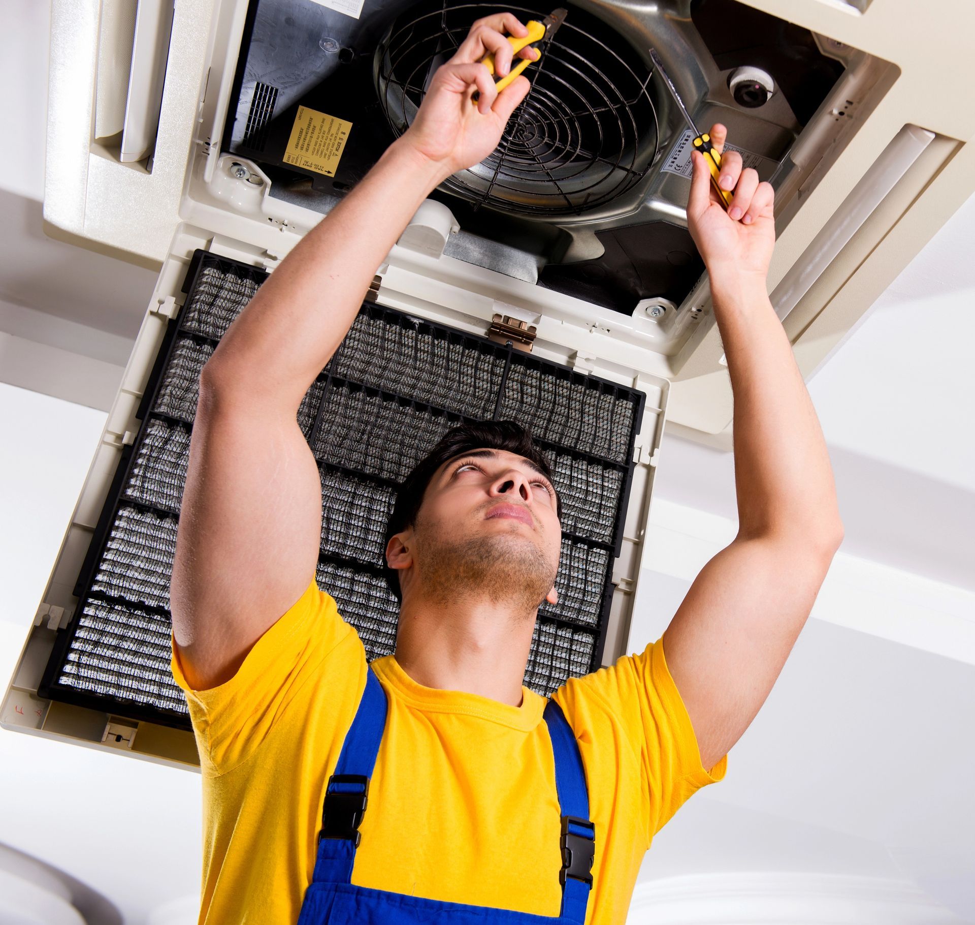 Technician in yellow shirt and blue overalls servicing a ceiling air conditioner unit