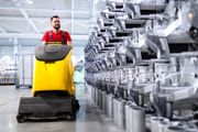 Un operaio guida una lavasciuga pavimenti gialla in un luminoso magazzino industriale, accanto a file di pezzi metallici.
