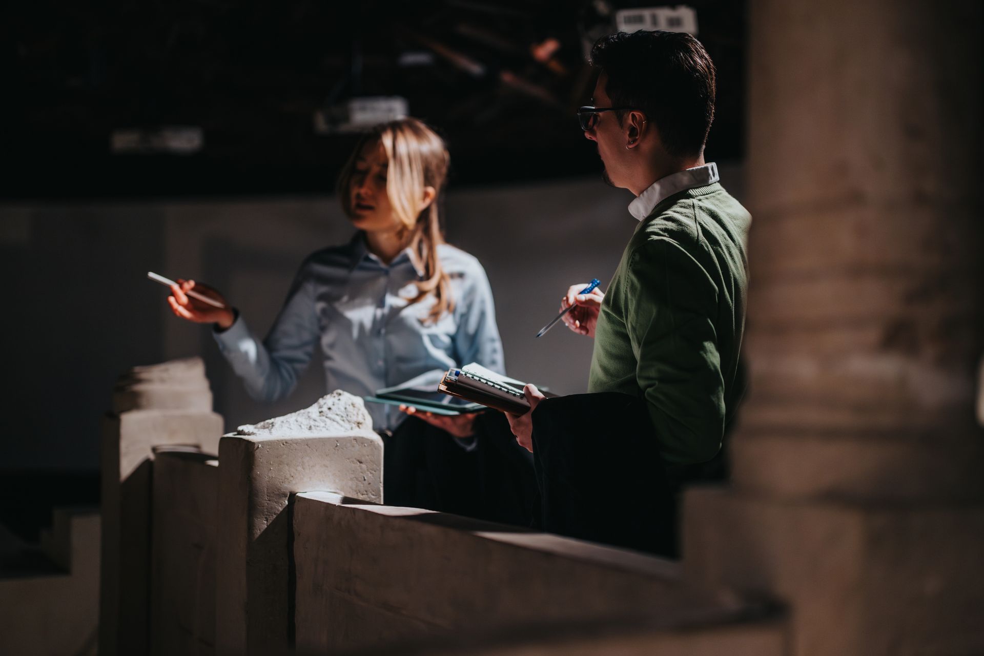 Two people talking in a dimly lit room, one gesturing while holding papers and the other listening.