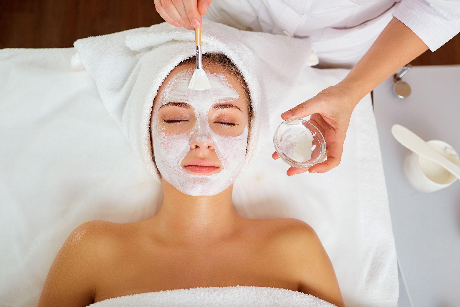 Mujer recibiendo un tratamiento facial en un spa. Una persona aplica una mascarilla blanca con una brocha.