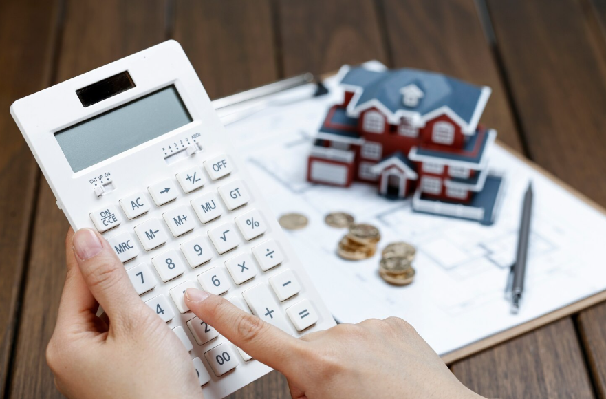 Manos usando una calculadora junto a una maqueta de una casa, papeles y monedas sobre un escritorio.