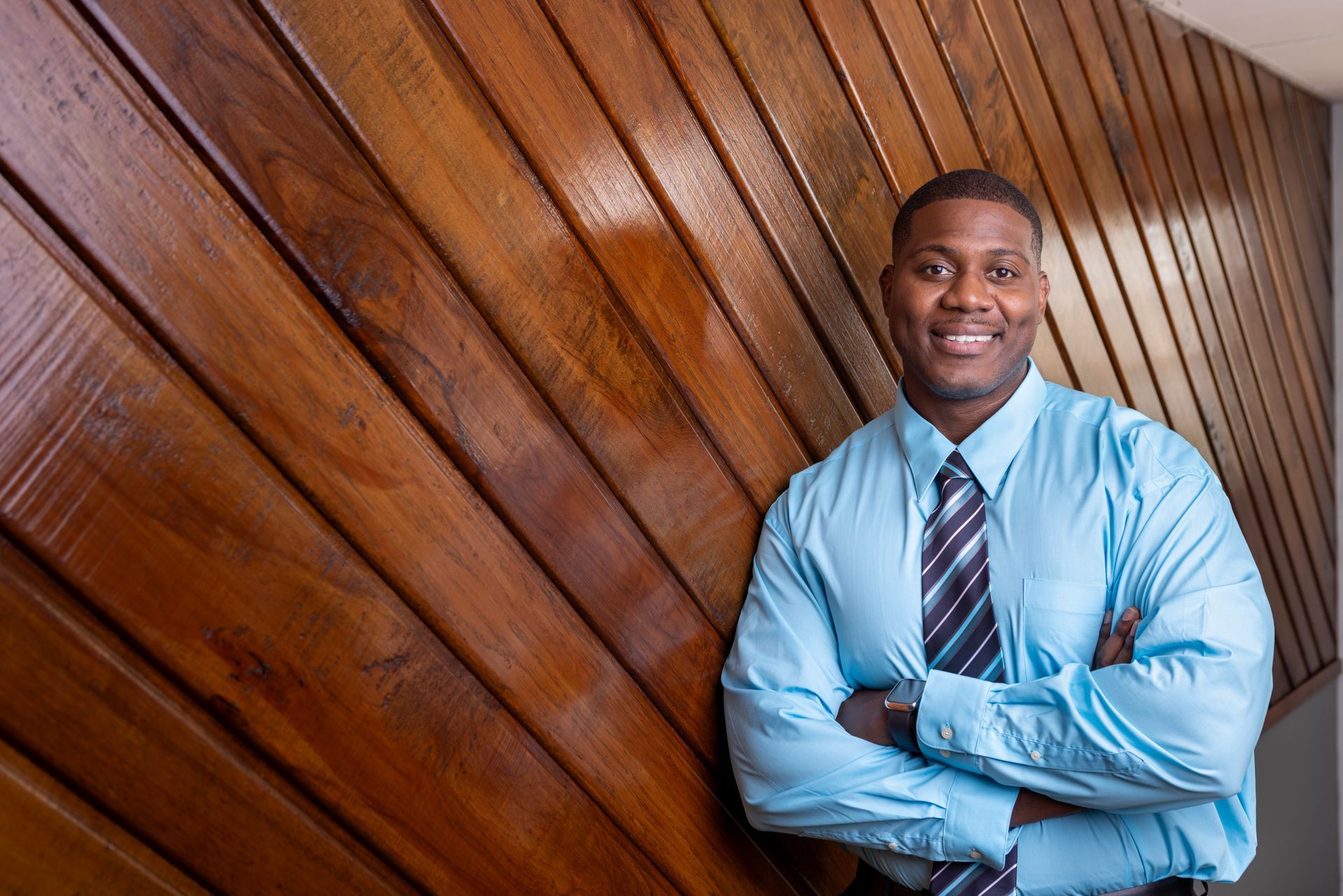 A man in a blue shirt and tie is standing in front of a wooden wall.