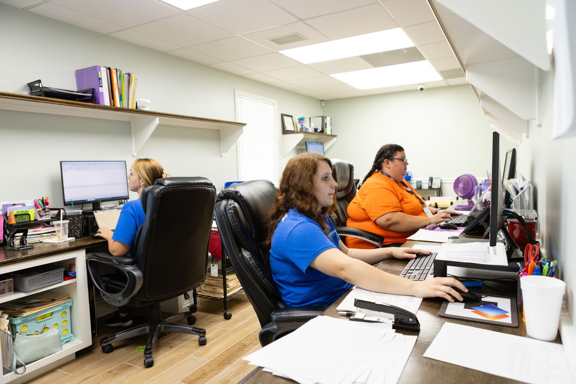 Three women are sitting at desks in an office working on computers.