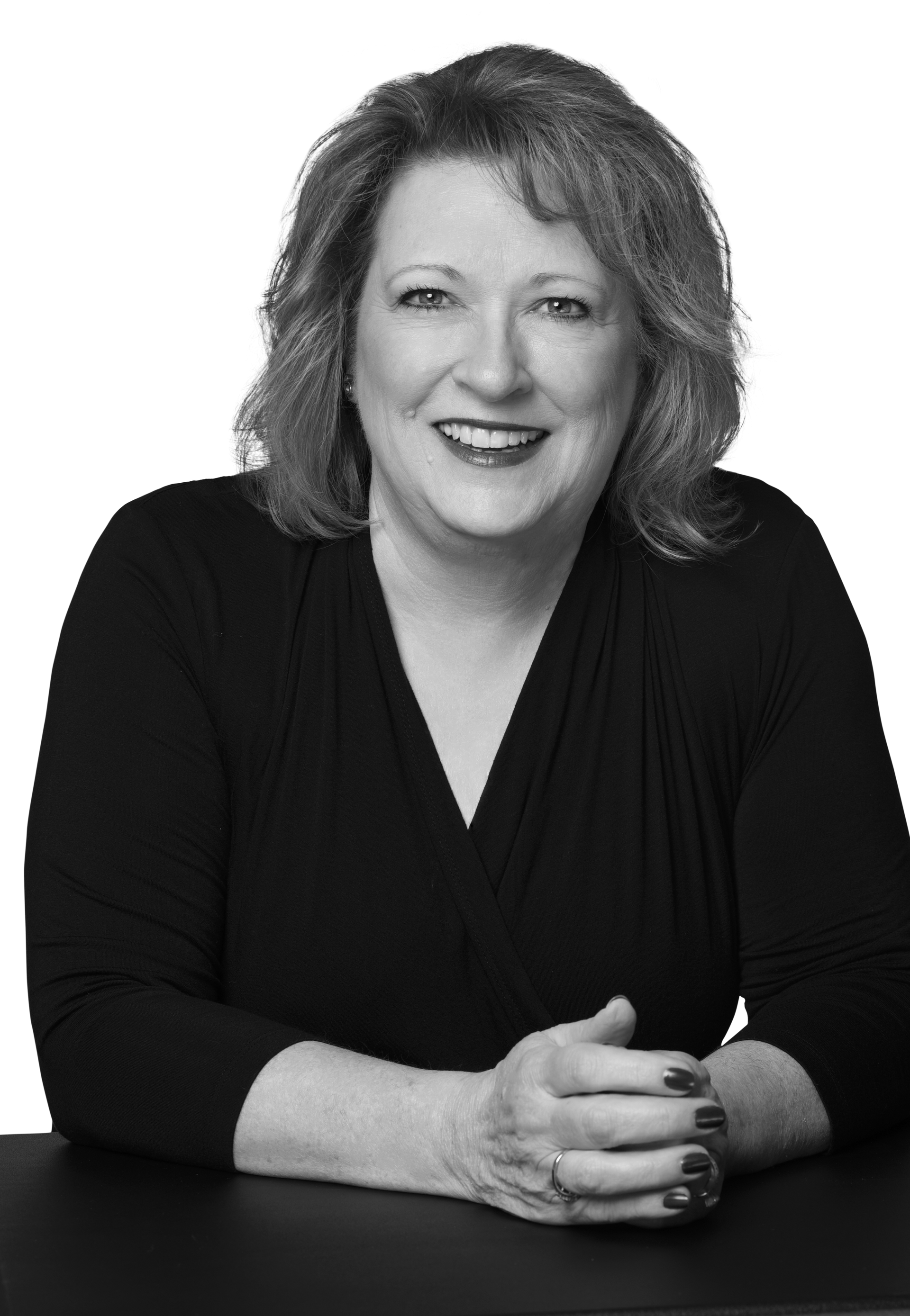 A woman is sitting at a table with her hands folded and smiling.