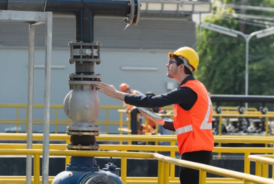 Um trabalhador com colete de segurança e capacete inspeciona uma válvula em um cano em um ambiente industrial.