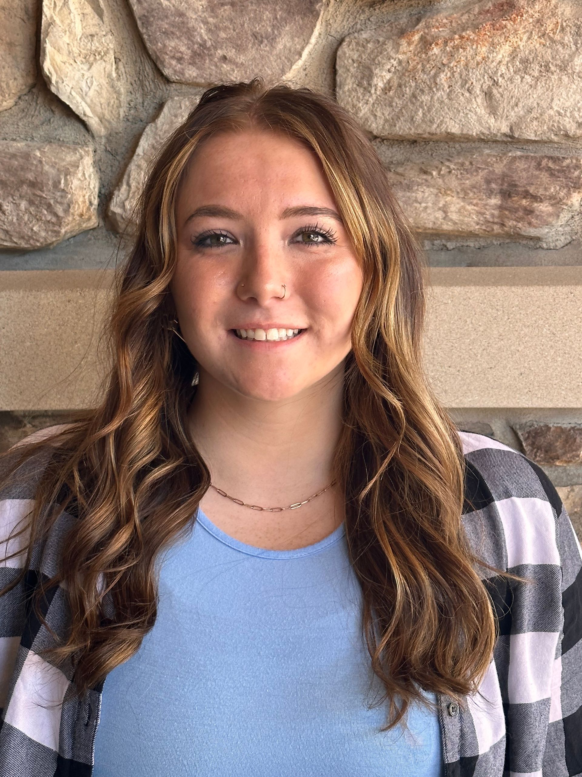 A young woman in a blue shirt and plaid jacket is smiling in front of a stone wall.