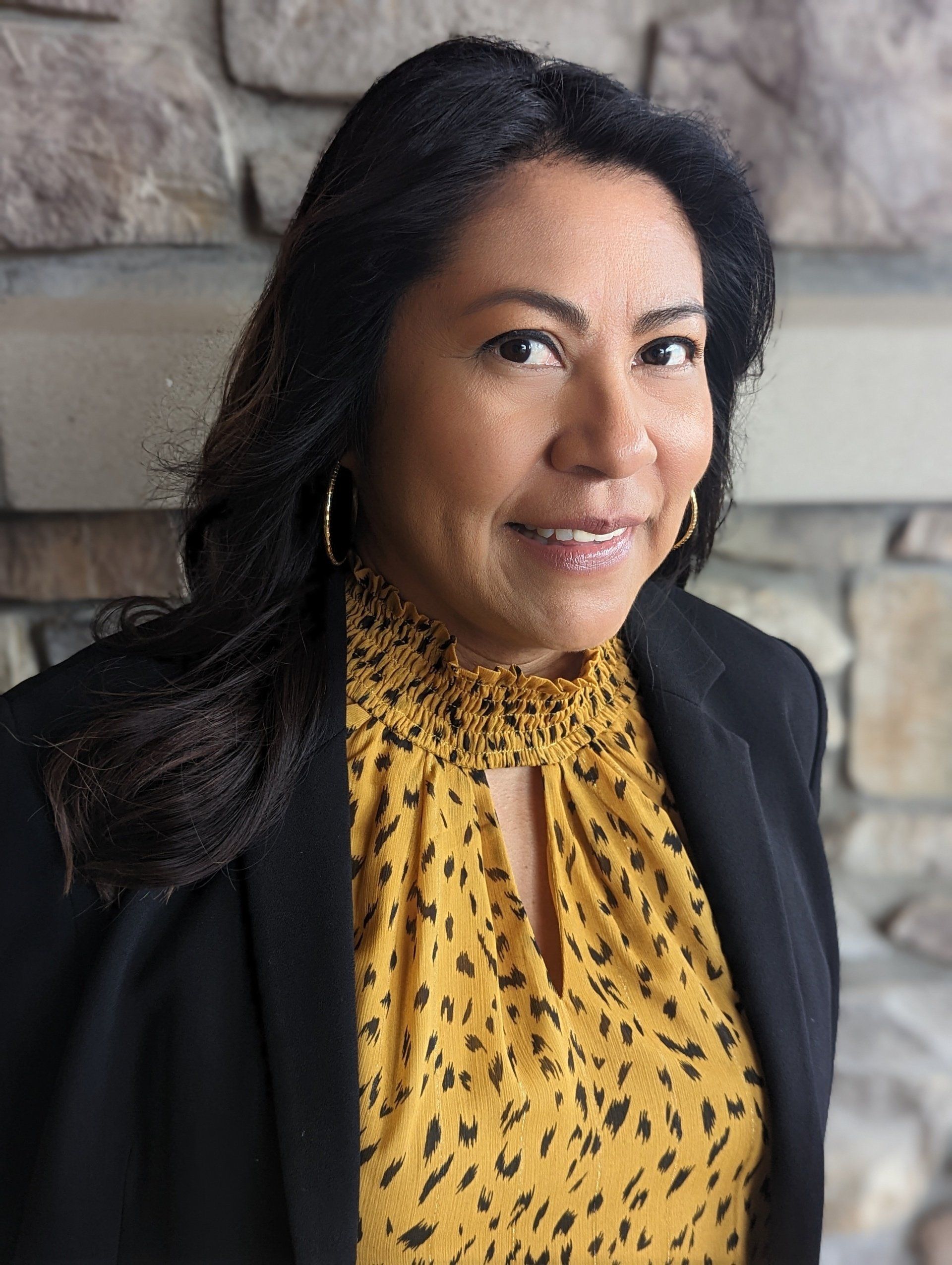 A woman wearing a yellow leopard print shirt and a black jacket is standing in front of a stone wall.