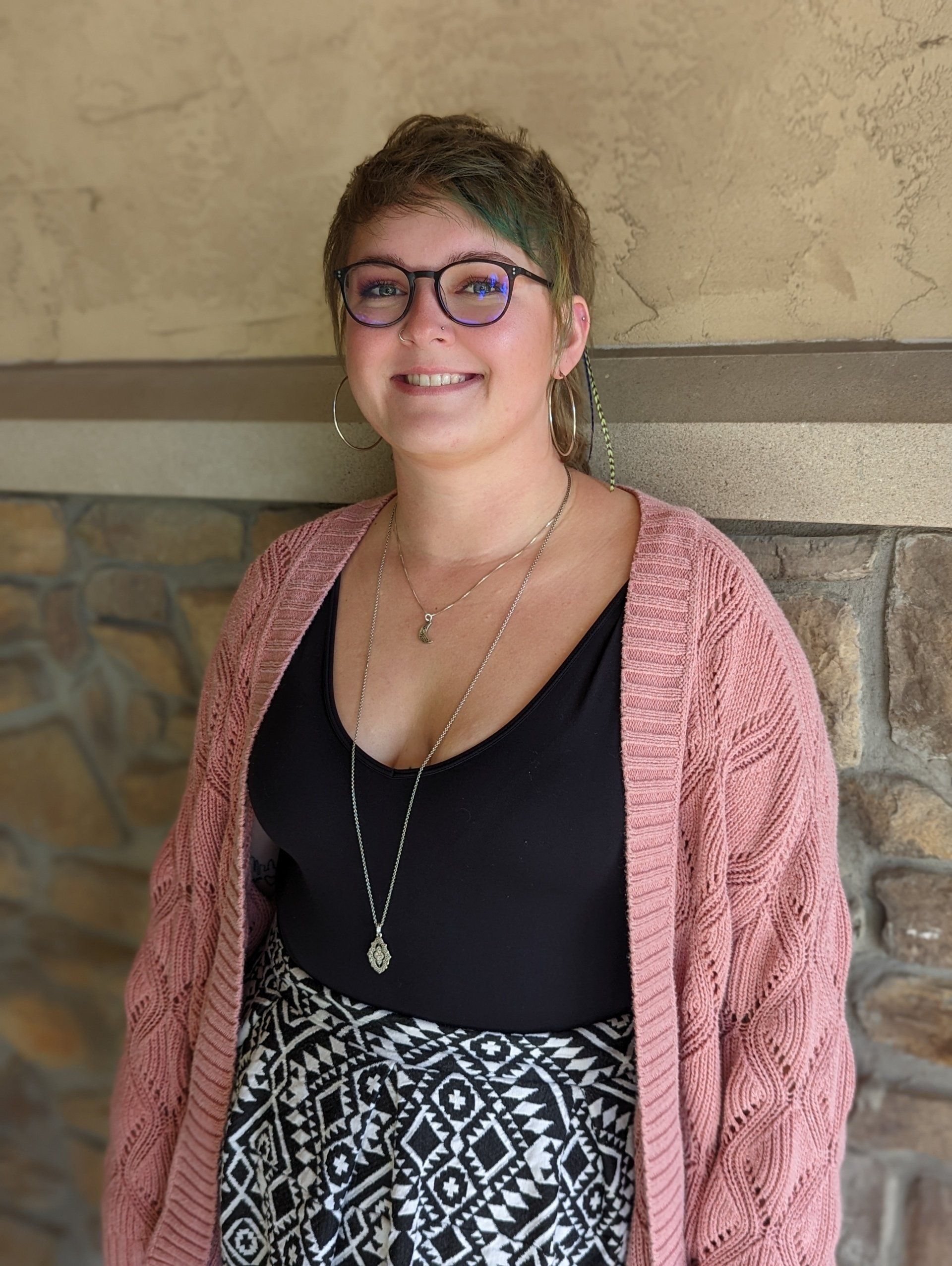A woman wearing glasses and a pink cardigan is standing in front of a stone wall.