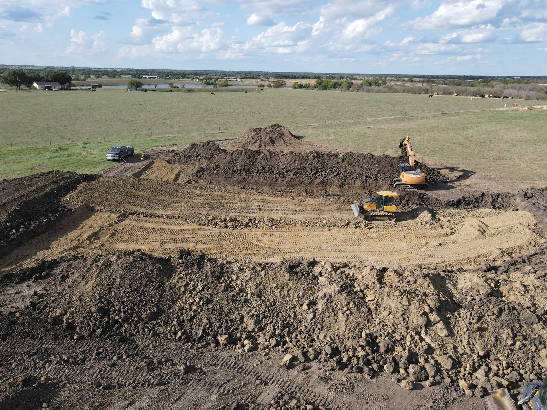 An aerial view of excavators working on a large, rectangular trench in a rural, open field under a blue sky.