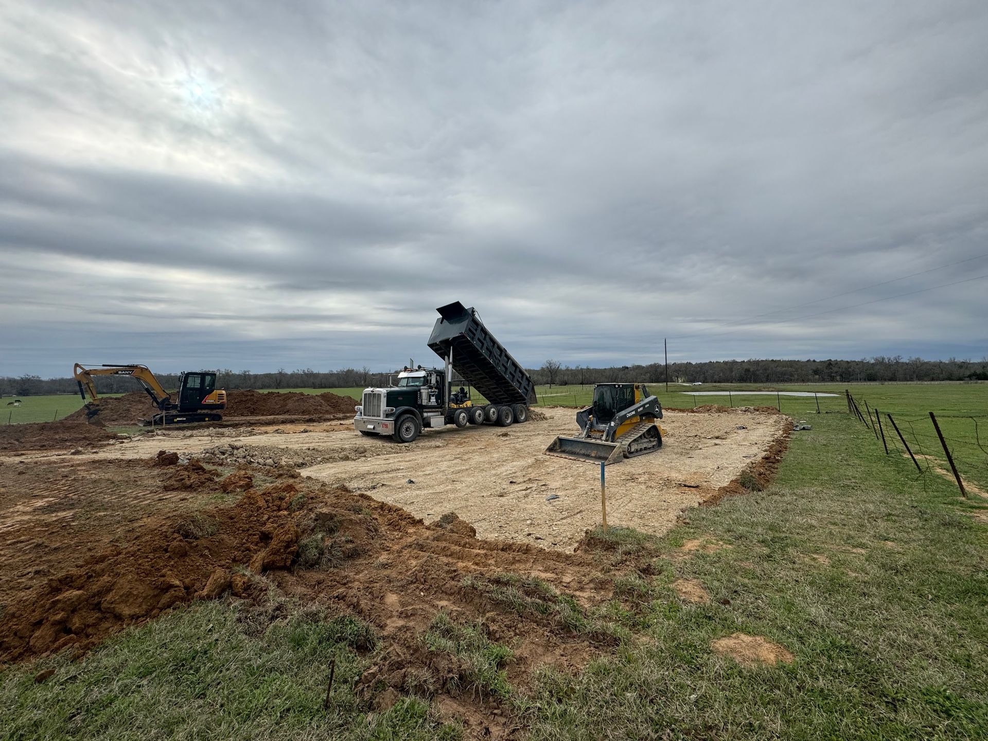 Construction equipment and a dump truck work on a dirt lot in a grassy field under a cloudy sky.
