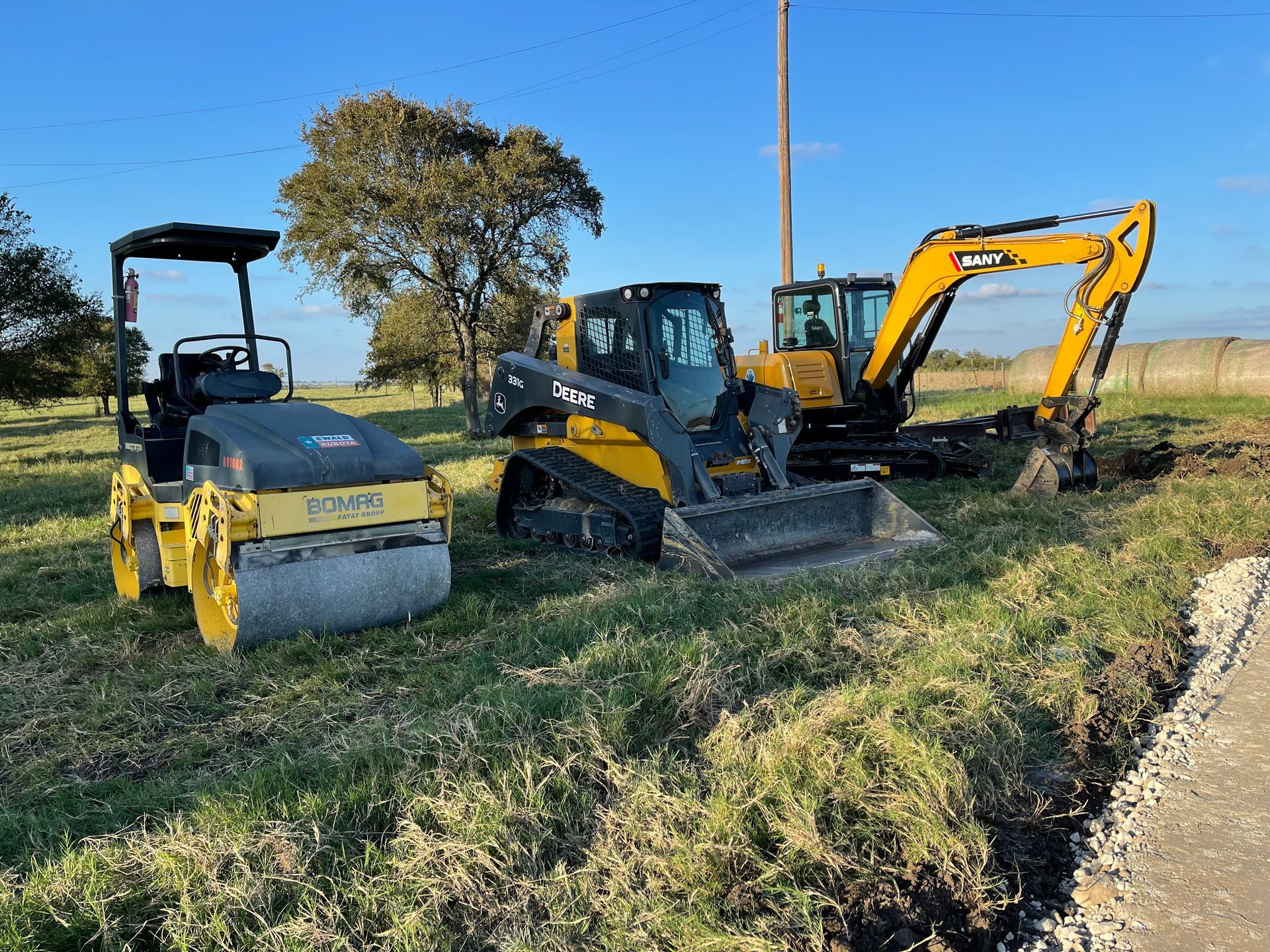 A yellow soil compactor, a yellow and black skid steer, and a yellow excavator parked in a grassy field.