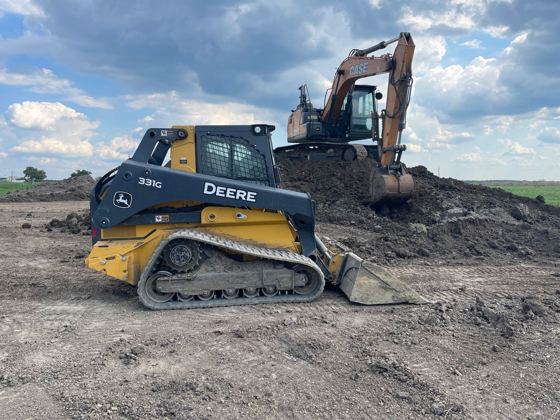 A yellow and black John Deere track loader sits on dirt with a large excavator working on a pile of soil behind it.