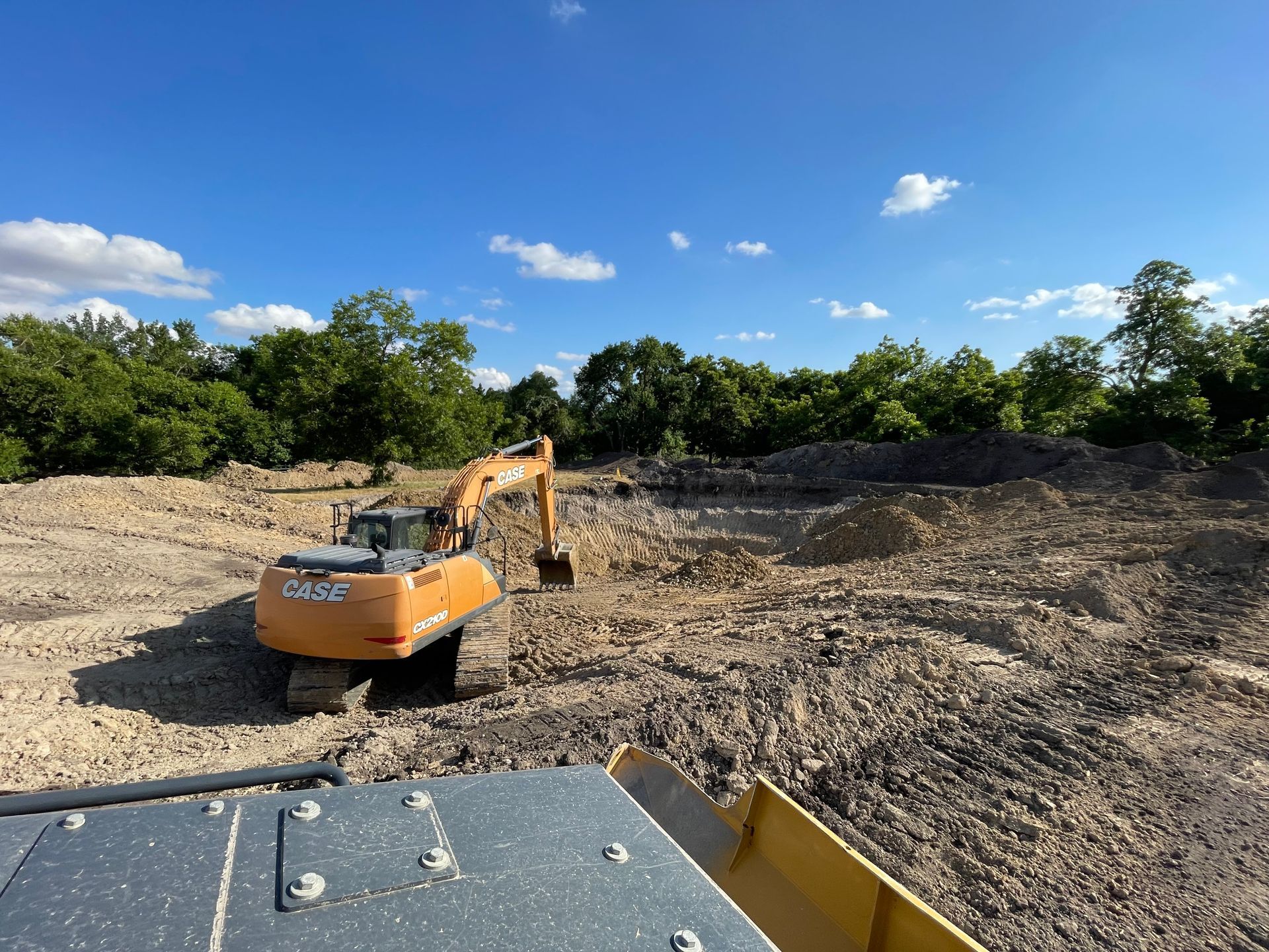 An orange excavator working in a large, rocky, uneven dirt clearing under a clear blue sky.