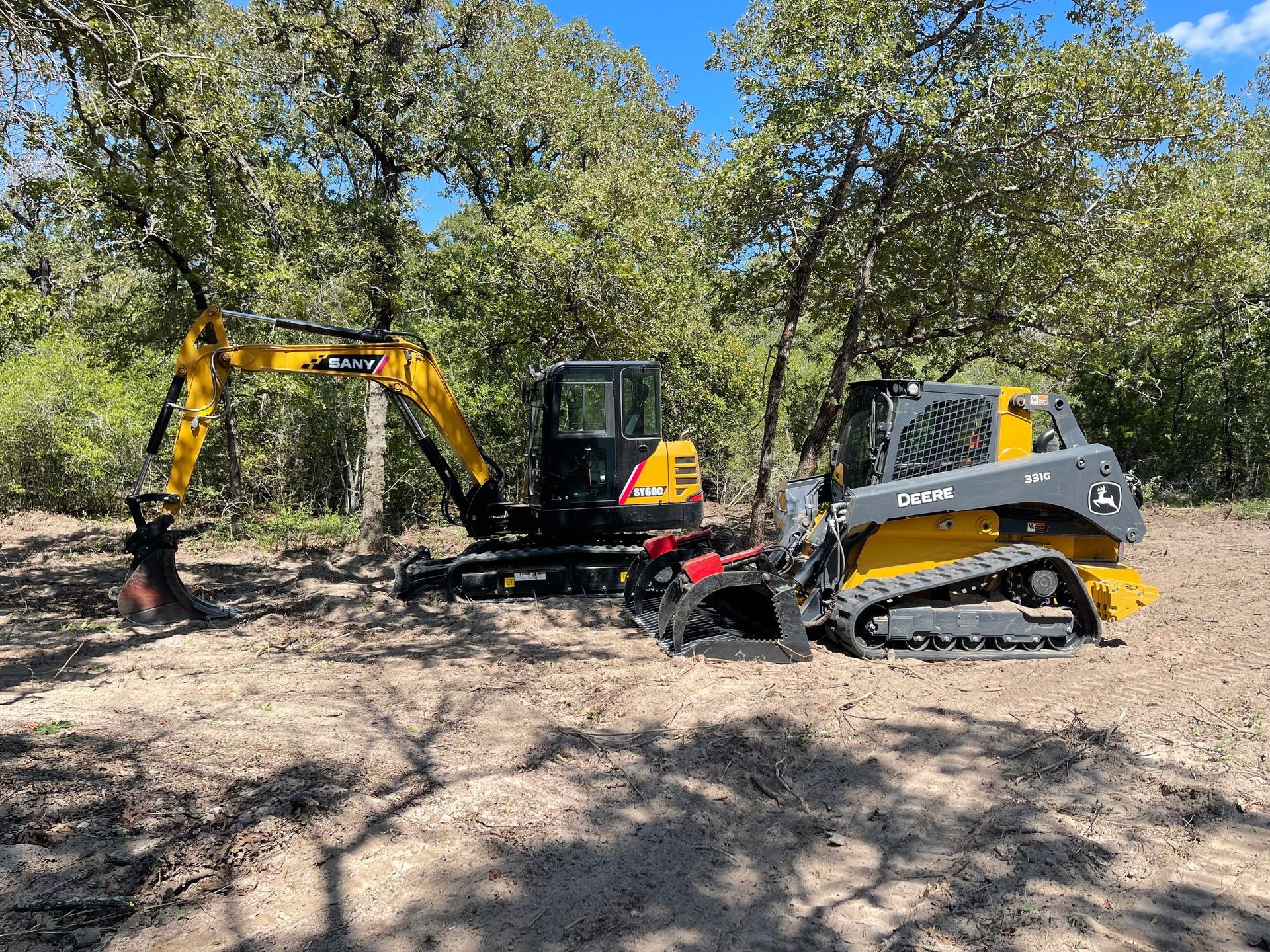 A yellow and black excavator and a yellow track loader parked in a wooded area on a sunny day.