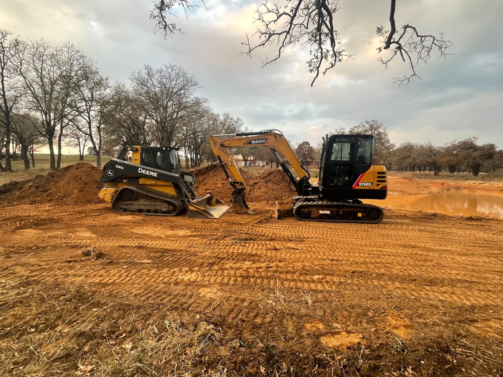 A compact excavator and a tracked skid steer parked on a dirt construction site under a cloudy sky.