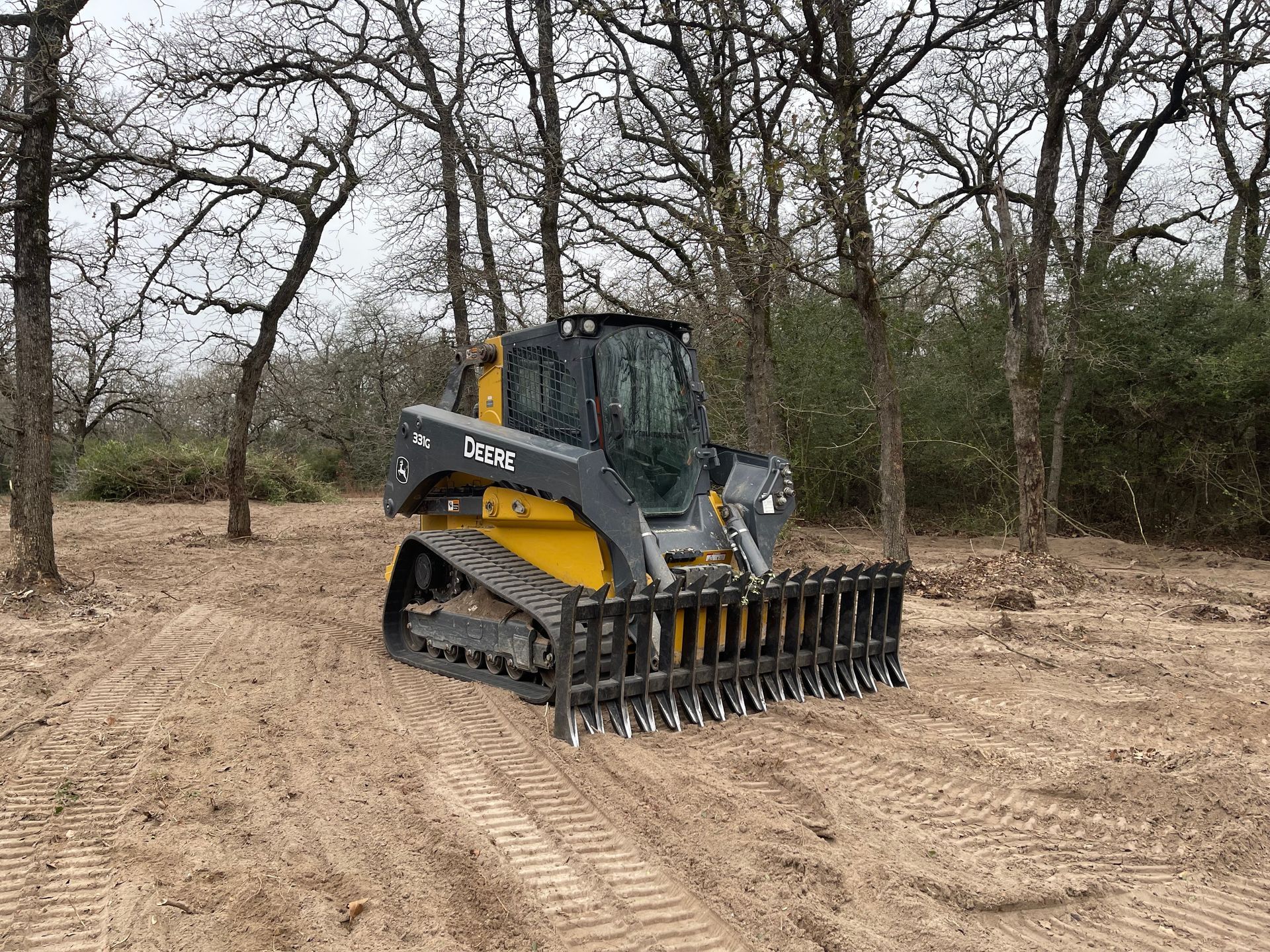 A yellow and gray Caterpillar compact track loader with a root rake attachment sits in a cleared, wooded area.