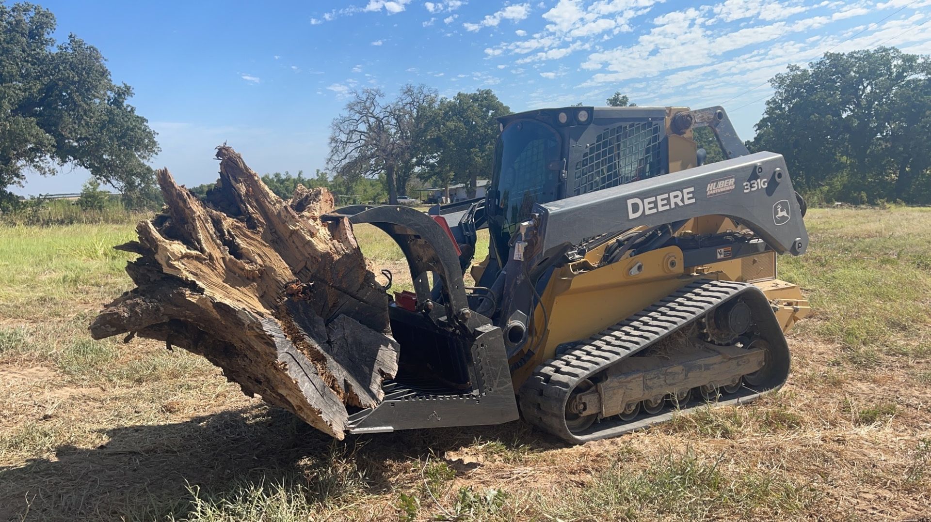 A yellow John Deere compact track loader with a tree grapple attachment carries a large, uprooted tree stump in a field.