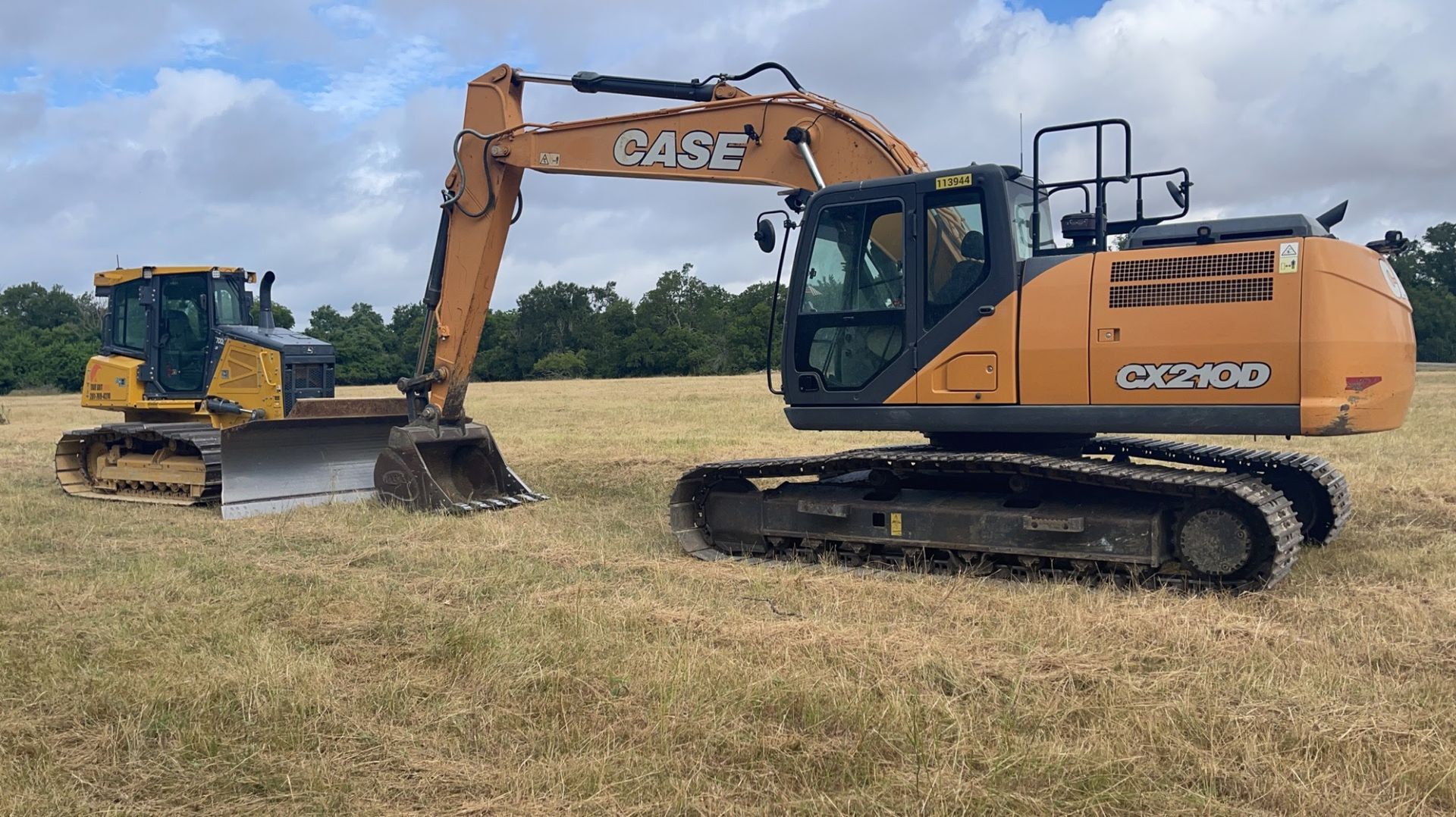 A tan Case crawler excavator and a yellow bulldozer parked on a grassy field under a cloudy sky.